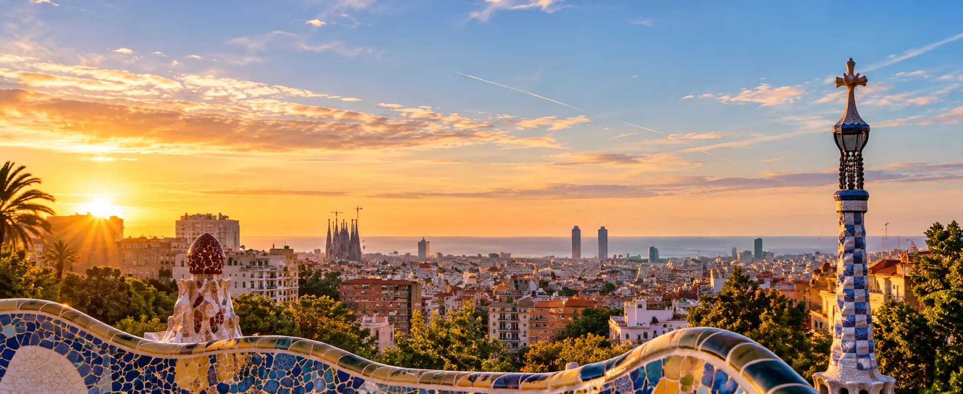 View of Barcelona from Park Guell at sunrise, with the mosaic bench in the foreground and the Sagrada Familia and the seafront towers on the skyline.