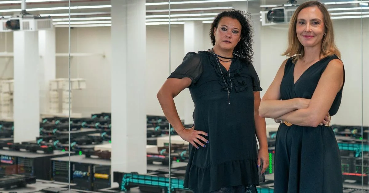 Maria Carolina Florian, left, and Paula Petrone, right, both dressed in black, stand in front of a glass wall with rows of supercomputer racks behind it.