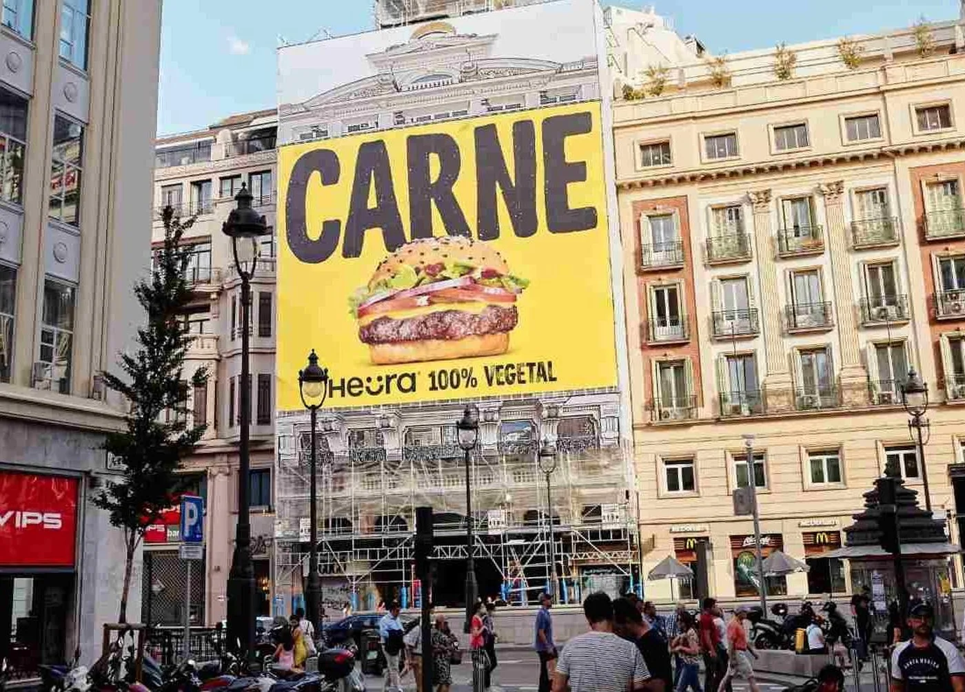 A large yellow Heura billboard reading CARNE above a burger and the words Heura 100% vegetal, on the facade of a building over a busy city street.