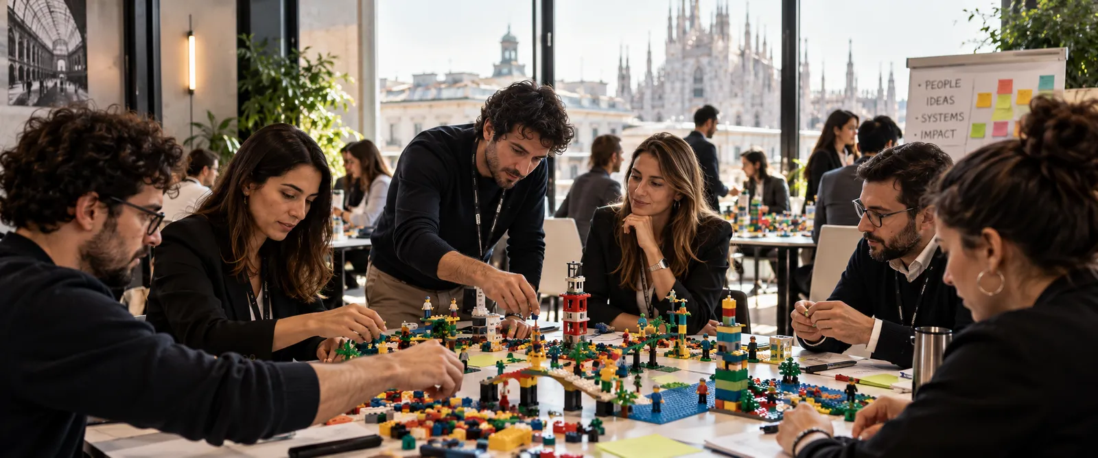 A workshop group building models with coloured bricks around a table, with Milan cathedral through the window.