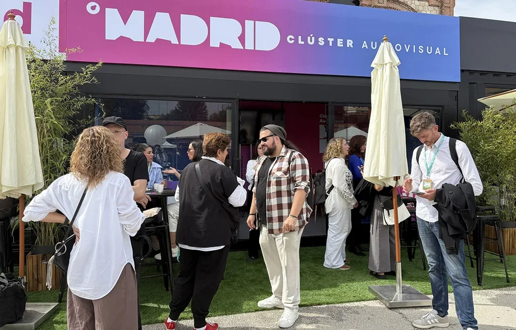 People chat on artificial grass in front of a black container pavilion with a pink and blue banner reading Madrid Clúster Audiovisual, between white parasols.