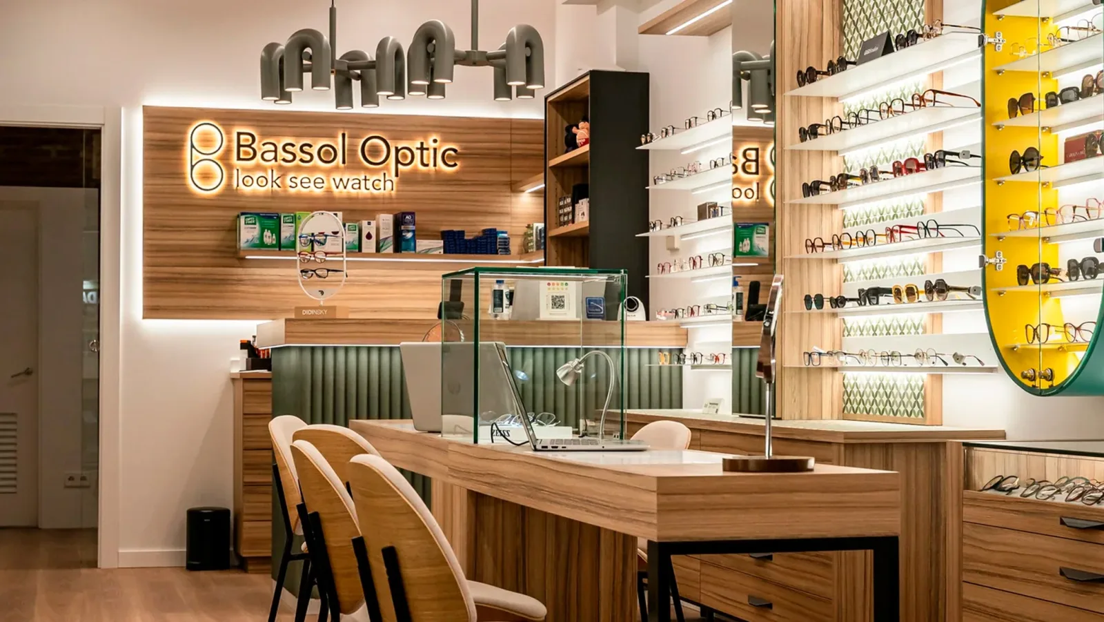Inside a Bassol Optic shop: an illuminated sign reading Bassol Optic, look see watch, over a wooden counter, with shelves of glasses and sunglasses on the right.